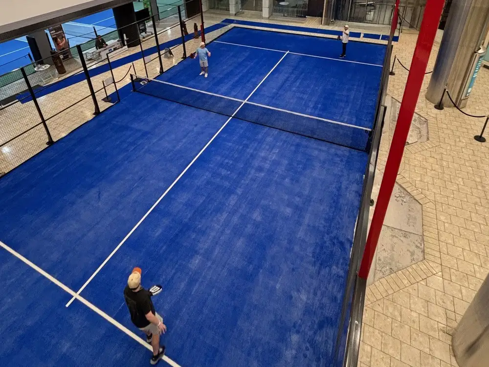 Players mid-game on Pickleball America's blue padel court, seen from above, with its glass walls and the Stamford Town Center mall floor alongside.