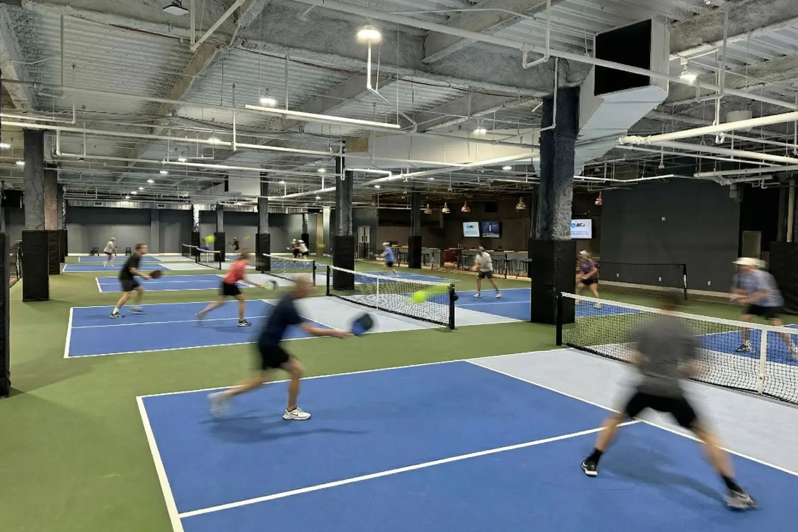 Wide view across the main floor: rows of blue pickleball courts separated by green run-off, players mid-rally on several of them, under an exposed ceiling of ductwork and pipes.